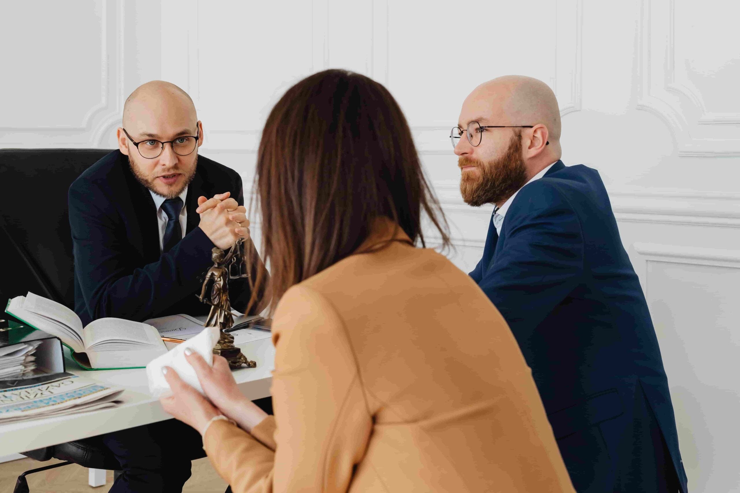 Three people in business attire sit at a desk having a discussion, with documents, books, and a small statue of Lady Justice visible on the table. Their focused conversation suggests they are reviewing important matters—perhaps weighing what should be considered before agreeing to a severance package in New Jersey.