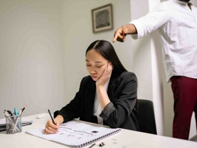 A woman sits at a desk writing in a notebook, looking down. A standing person behind her points a finger toward her, creating an atmosphere that raises the important question: What Steps Should I Take to Prove Age Discrimination at Work in New Jersey?.