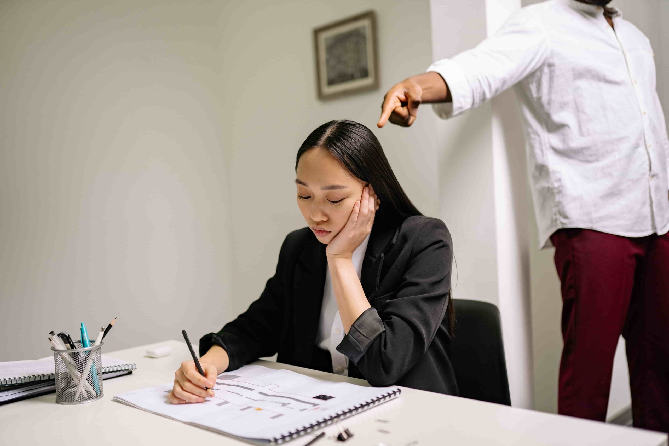 A woman sits at a desk writing in a notebook, looking down. A standing person behind her points a finger toward her, creating an atmosphere that raises the important question: What Steps Should I Take to Prove Age Discrimination at Work in New Jersey?.