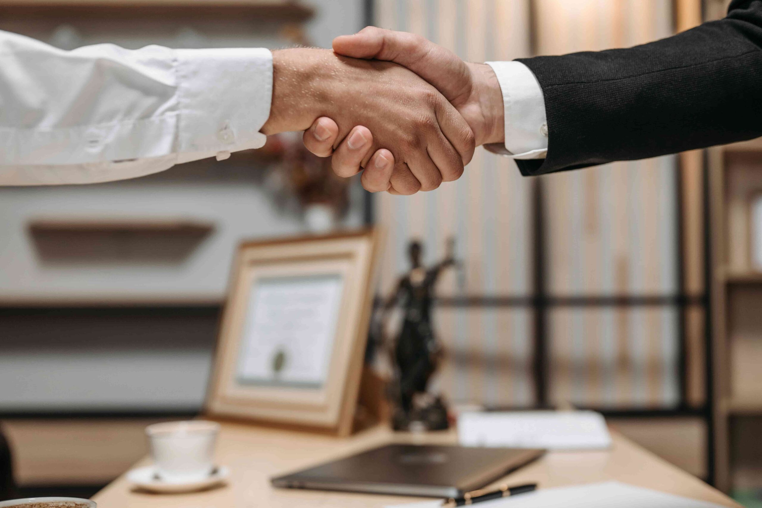 Two people are shaking hands over a desk with a cup, folder, and framed certificate in the background, possibly discussing what type of lawyer handles discrimination cases in New Jersey.