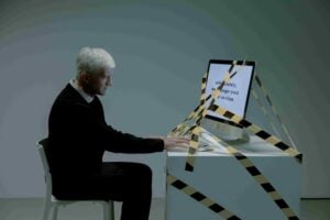 An older man sits at a desk typing on a computer monitor wrapped in caution tape, with unreadable text on the screen, in a sparsely lit room—a visual metaphor that raises questions such as, “Which Hiring Practices May Be Considered Age Discrimination in New Jersey?”.
