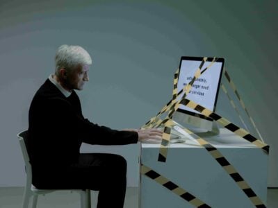 An older man sits at a desk typing on a computer monitor wrapped in caution tape, with unreadable text on the screen, in a sparsely lit room—a visual metaphor that raises questions such as, “Which Hiring Practices May Be Considered Age Discrimination in New Jersey?”.