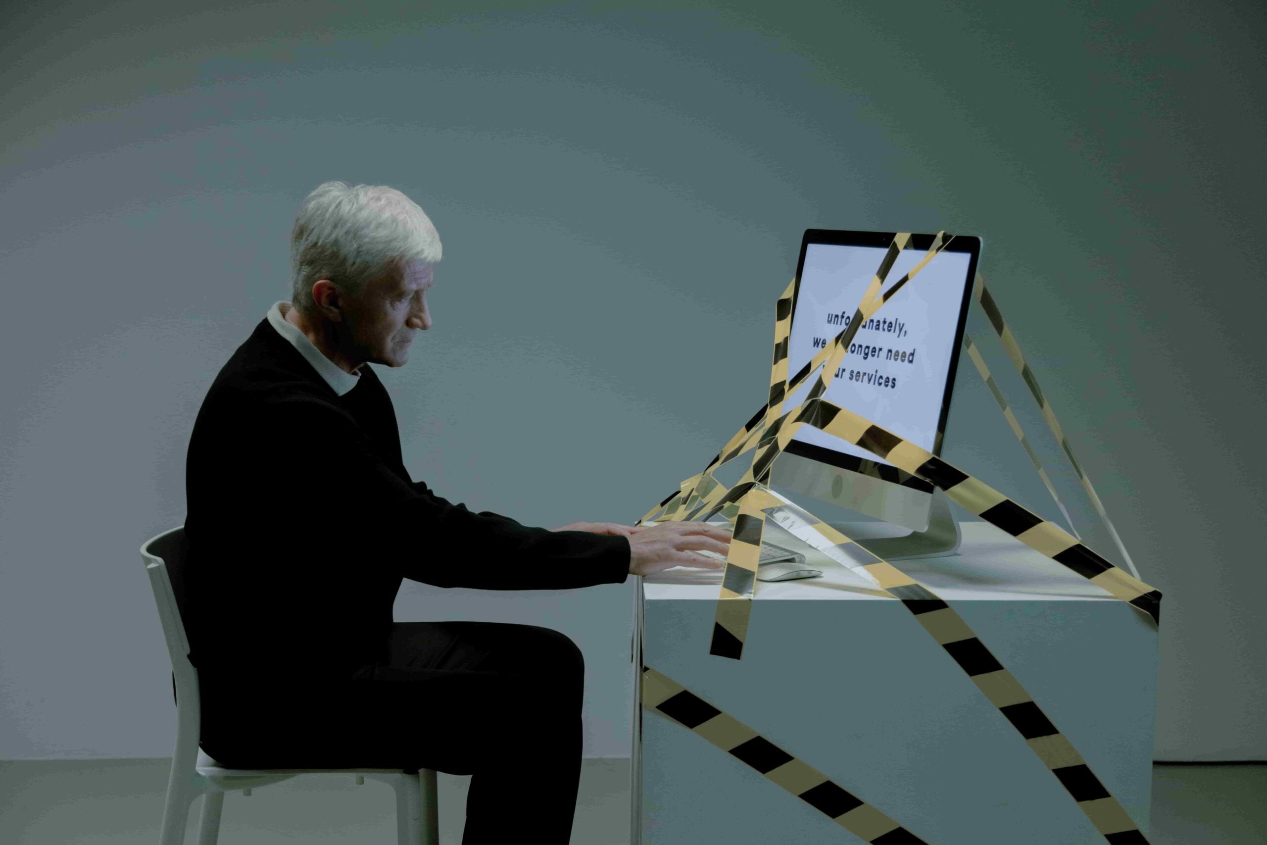 An older man sits at a desk typing on a computer monitor wrapped in caution tape, with unreadable text on the screen, in a sparsely lit room—a visual metaphor that raises questions such as, “Which Hiring Practices May Be Considered Age Discrimination in New Jersey?”.