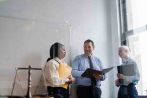 Three professionals in business attire stand by a window, discussing documents in an office with a whiteboard and legal scale visible, as they consider how Whistleblower Protections Work in New Jersey.