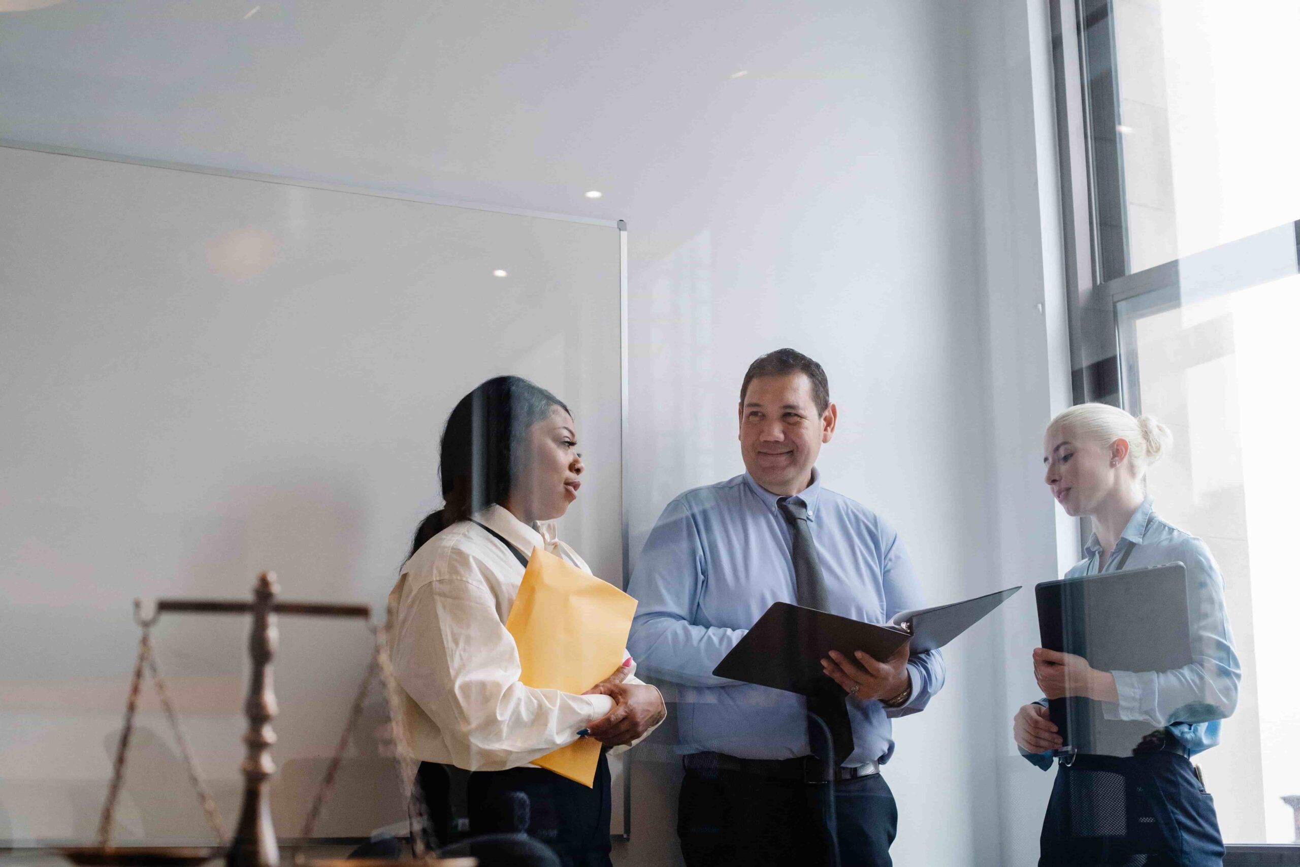 Three professionals in business attire stand by a window, discussing documents in an office with a whiteboard and legal scale visible, as they consider how Whistleblower Protections Work in New Jersey.