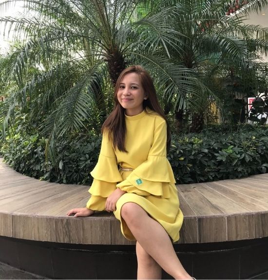 A woman in a yellow dress sits on a curved wooden bench in front of lush green plants and palm trees, smiling at the camera outside Lacy Employment Law Firm.