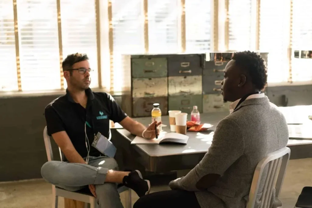 Two men sit across from each other at a table in a sunlit room, engaged in conversation about how to resolve wrongful termination. There are drinks, papers, and a notebook on the table as they discuss the details and timeline of the situation.