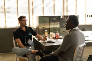 Two men sit across from each other at a table in a sunlit room, engaged in conversation about how to resolve wrongful termination. There are drinks, papers, and a notebook on the table as they discuss the details and timeline of the situation.