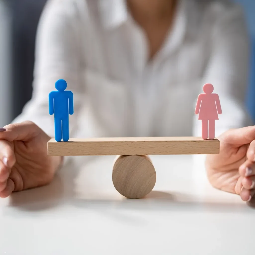 A person holds a wooden balance scale with blue male and pink female figures on opposite ends, symbolizing gender equality—an image that resonates with the mission of a Gender Discrimination Attorney.