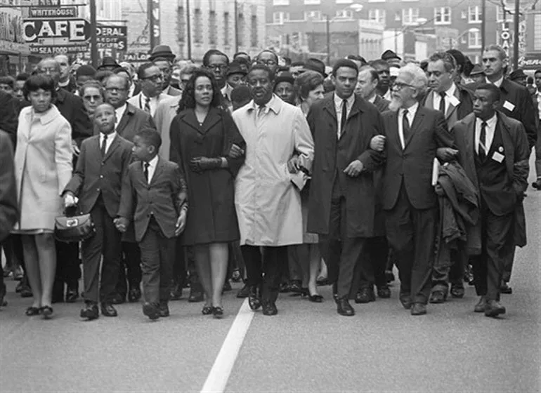 A diverse group of men, women, and children, guided by a Civil Rights Attorney, march arm in arm on a city street during a civil rights demonstration.