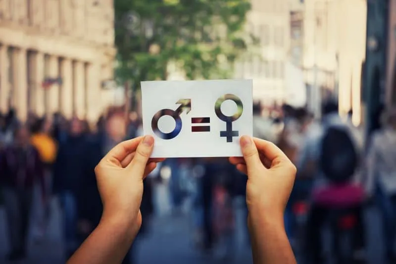 Two hands hold a paper with male and female gender symbols connected by an equals sign, highlighting equality—a powerful image for a Gender Discrimination Attorney—set against a blurred city street and crowd in the background.