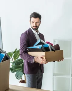 A man in a suit holds a cardboard box filled with office supplies, standing in an office setting near a plant and empty shelves—an image that suggests the impact of harassment or a hostile work environment.