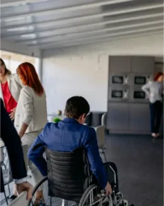 A person in a wheelchair is seated at a table in a modern office space, with three others standing and talking in the background, highlighting the importance of disability rights and inclusion in the workplace.
