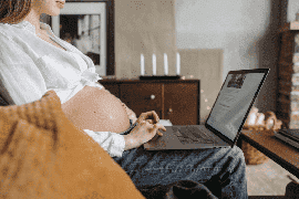 Pregnant person sitting on a couch using a laptop, their belly visible, possibly searching for Philadelphia pregnancy discrimination lawyers in the comfort of their living room.