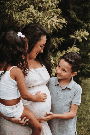 A pregnant woman stands outdoors, smiling as a young girl hugs her and a young boy touches her baby bump—a moment that highlights the importance of support and awareness, values upheld by Philadelphia pregnancy discrimination lawyers.