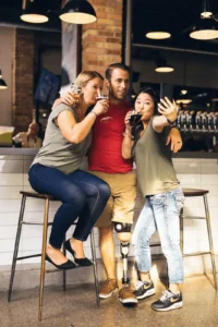 Three people pose for a photo at a bar, reminiscent of relaxed NJ Workplaces camaraderie. One woman sits on a stool, another stands taking a selfie, and a man with a prosthetic leg stands between them. All are smiling and holding drinks.