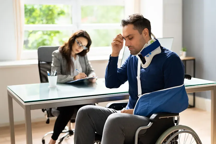 A man in a wheelchair with a neck brace and arm sling sits with eyes closed, looking distressed, while a woman from a team of personal injury lawyers takes notes at her desk in an office.