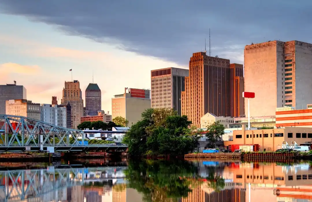 Cityscape with tall buildings and a bridge reflected in a calm river under a cloudy sky. Dense greenery is visible near the water’s edge, capturing the tranquil beauty often found in New Jersey, where accessible design and reasonable accommodations are increasingly integrated into urban environments.