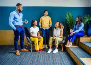 Five people in business casual attire are talking and smiling in a modern office space with blue and green decor, reflecting positive workplace communication.