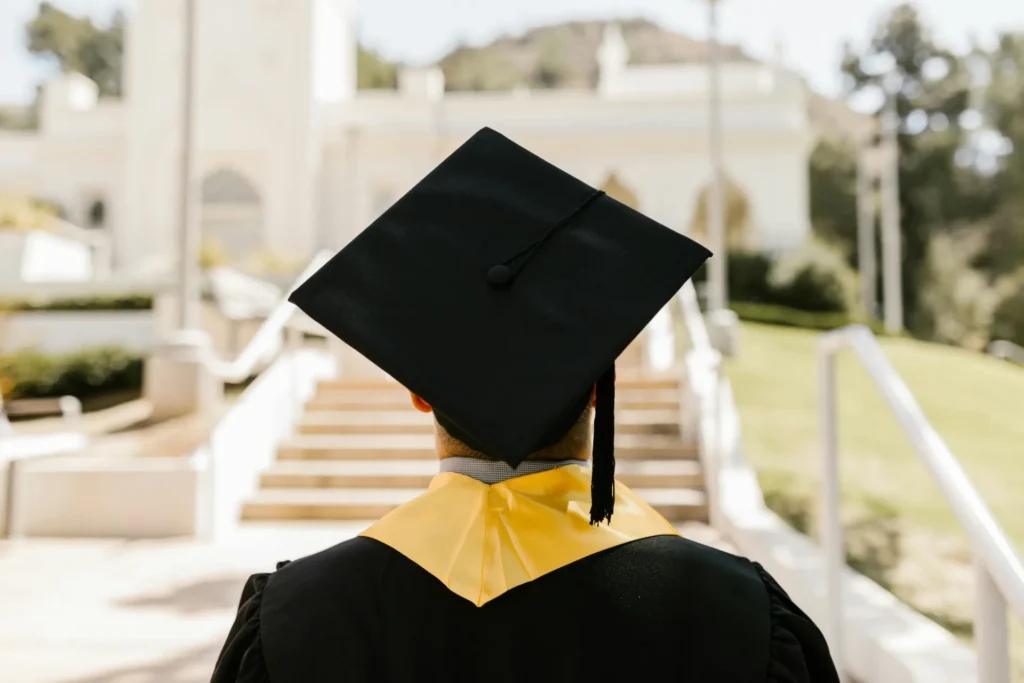 A graduate in a cap and gown stands facing outdoor steps, with a white building and greenery visible in the background, ready to embark on their professional journey—perhaps at places like Lacy Employment Law Firm.