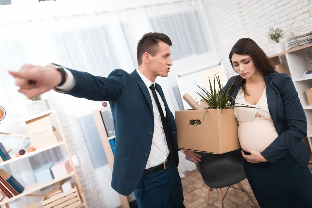 A man in a suit points toward the door while a pregnant woman holding a box of office items looks down sadly, highlighting the need for a Pregnancy Discrimination Lawyer.