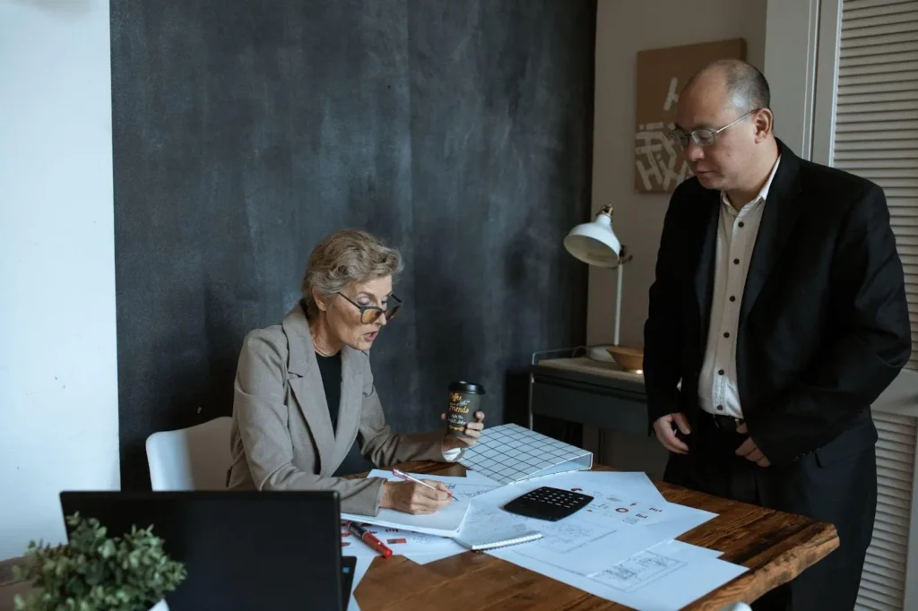Two people in business attire review documents at a desk with papers, a calculator, and a laptop in an office setting.