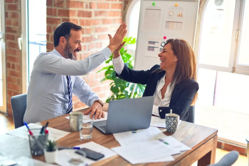 Two businesspeople sitting at a desk high-five each other, smiling, with papers, a laptop, and drinks in front of them in a modern office setting.