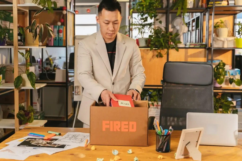 A person in an office packs personal items into a box labeled “Fired”; papers, pens, and plants are visible on the desk and shelves in the background.
