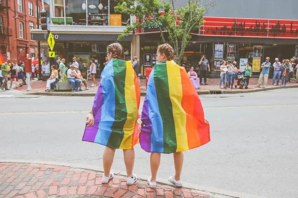 Two people wearing rainbow pride flags stand on a sidewalk facing a street, with bystanders and buildings visible in the background—an image that captures both the vibrancy of Pride and the ongoing need for support from an LGBT discrimination lawyer.