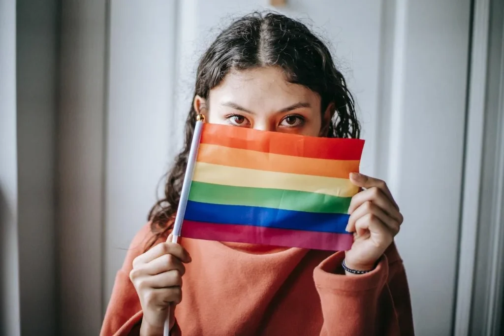 Person in an orange sweater holds a small rainbow pride flag in front of their face, with only their eyes visible, indoors against a light background—capturing the spirit and resilience that an LGBT discrimination lawyer proudly advocates for.
