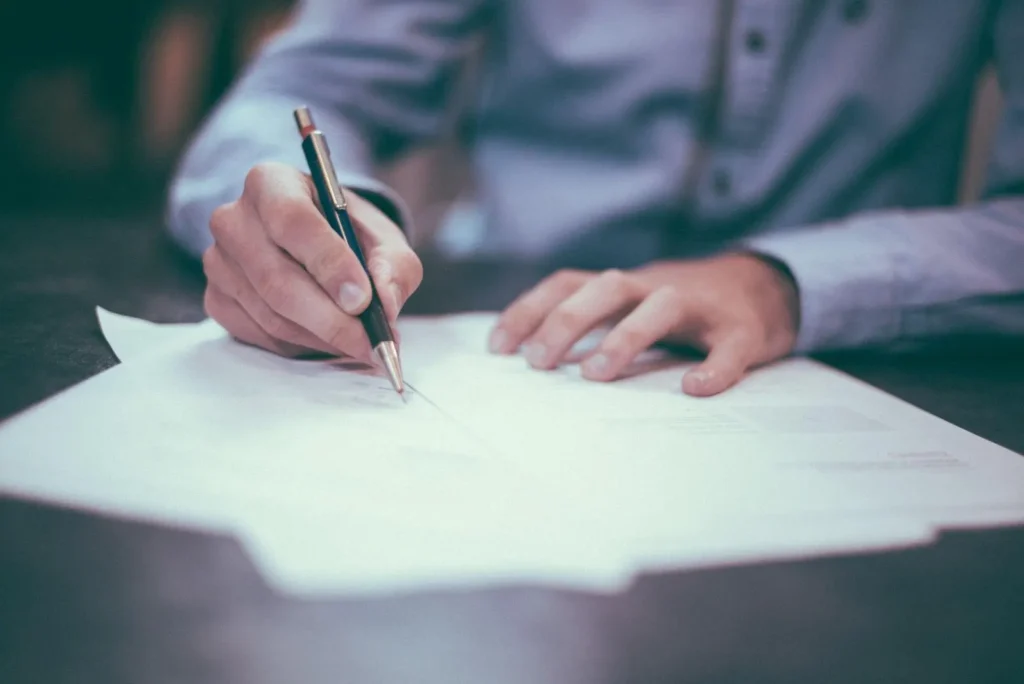 A person in a dress shirt writes with a pen on several sheets of paper spread out on a desk.
