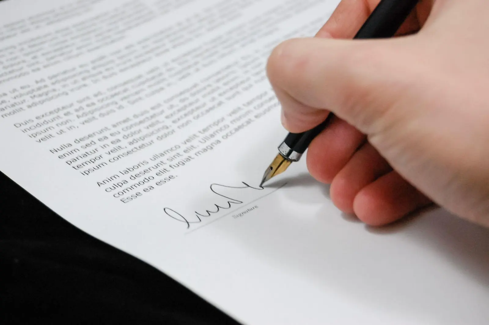 A hand holding a pen signs a document at the bottom, where there is a signature line and printed text above on the paper.