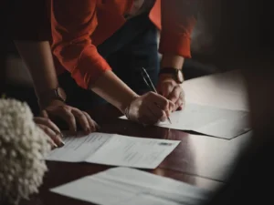 Person in a red jacket leans over a table, signing a document with a pen, while another person stands nearby. Several papers are spread out on the wooden tabletop.