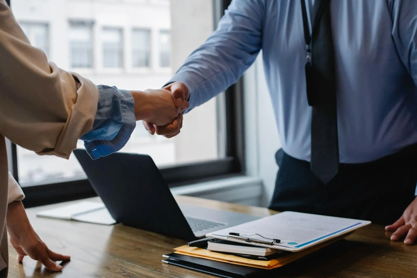 Two people in business attire shake hands across a desk with a laptop, clipboard, and documents, in an office with large windows.
