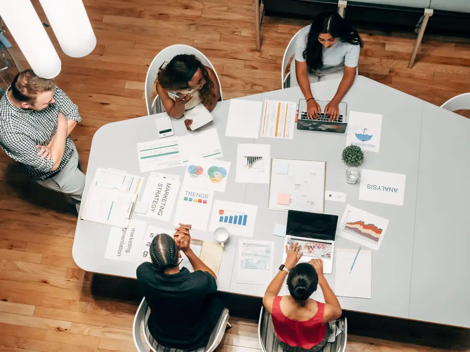 Five people sit around a table with laptops, charts, and documents, collaborating on business analytics and marketing trends in a modern office setting.