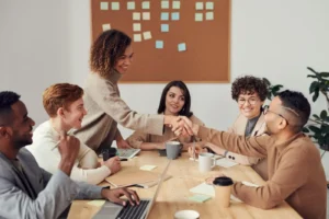 Six people sit around a table in a meeting room with coffee, laptops, and notes; two people are shaking hands while others watch and smile.