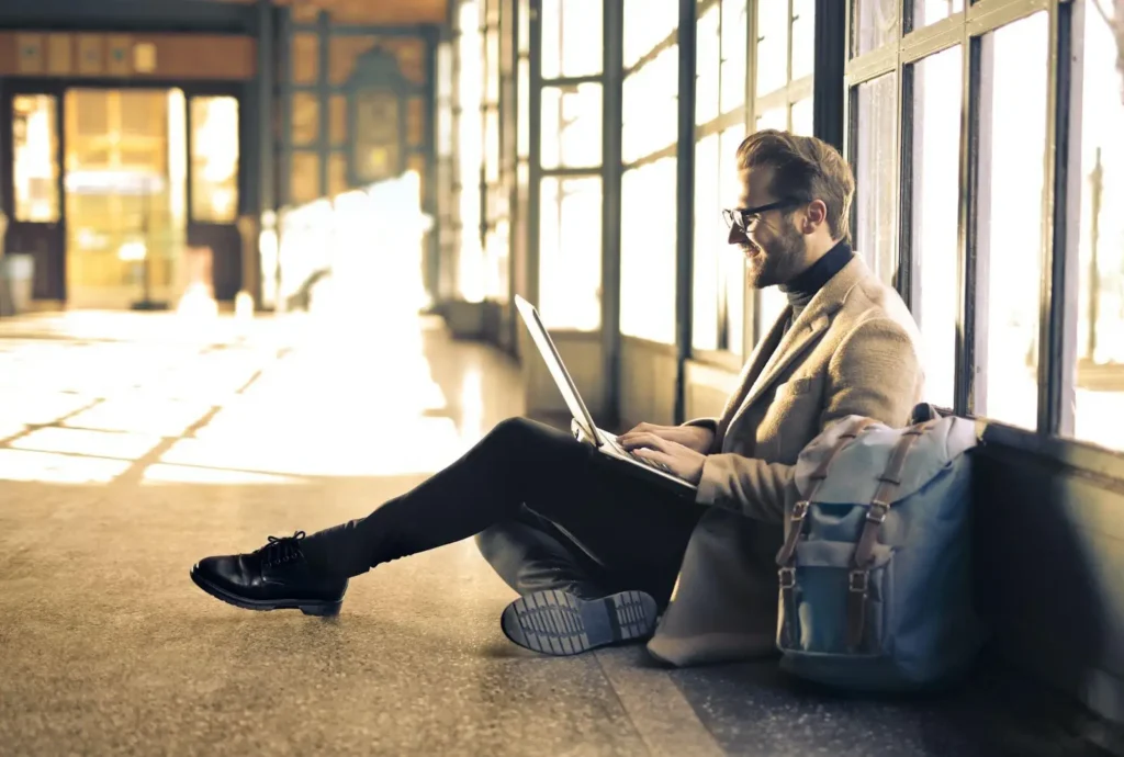 Man sitting on floor by large windows, typing on a laptop with a backpack next to him; sunlight streams in, illuminating the space—perhaps planning his next adventure and tracking FLSA Travel Time for work compliance.