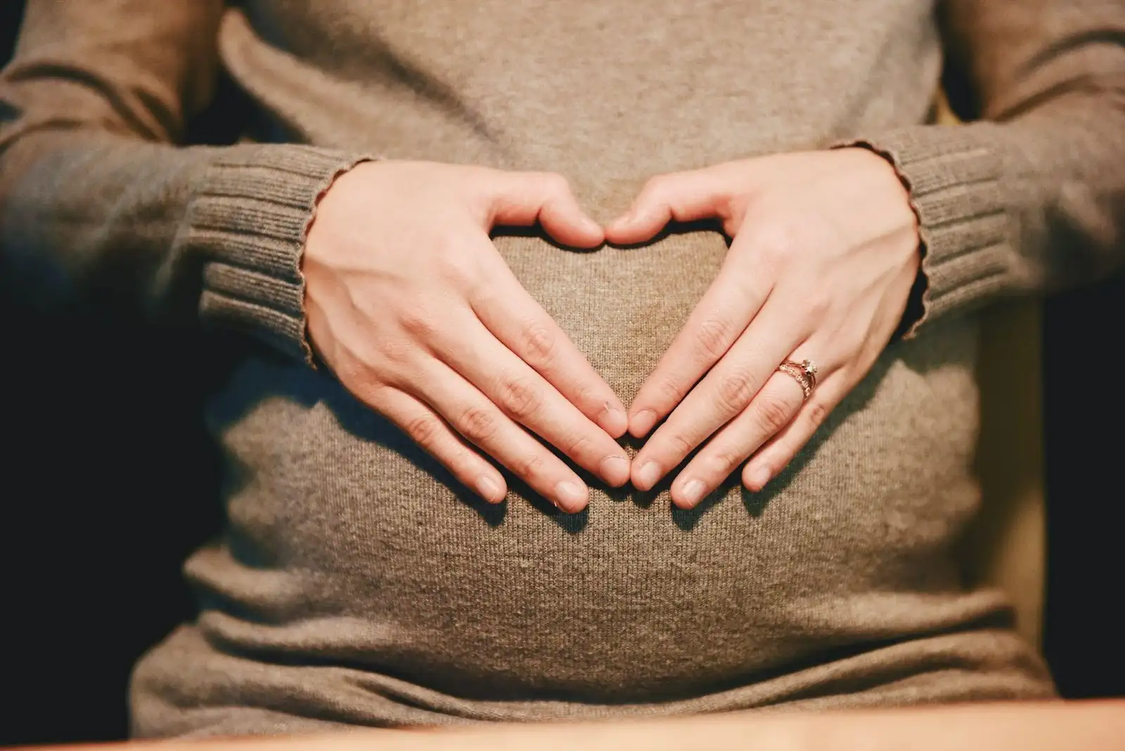 A person wearing a brown sweater forms a heart shape with their hands over their pregnant belly.