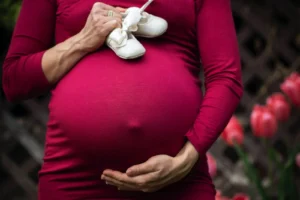 A person in a red dress holds white baby shoes and gently cradles their maternity belly, with blurred pink flowers in the background.