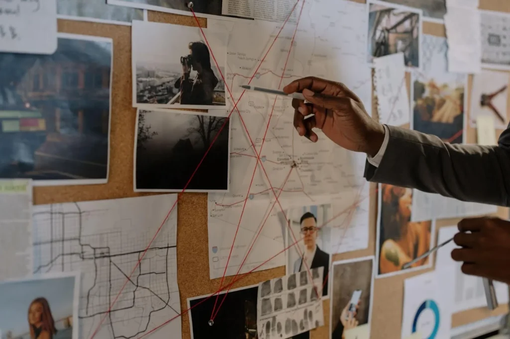 A person points a pen at a bulletin board covered with photos, maps, documents, and red string connecting various items in an investigative display—perhaps piecing together evidence for an EEOC case.