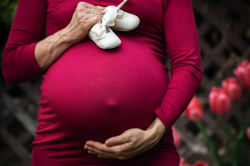 A person in a red maternity dress holds baby shoes over their pregnant belly with one hand and cradles the bump with the other. Pink flowers are visible in the background.