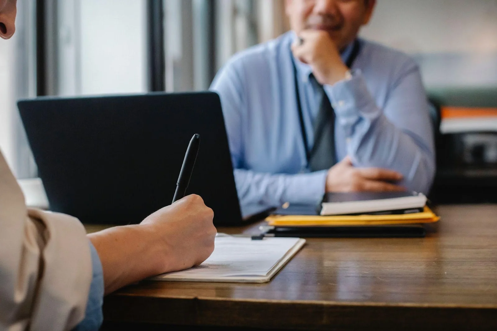 Two people sit at a desk; one writes in a notebook while the other sits across from them with a laptop and folders.