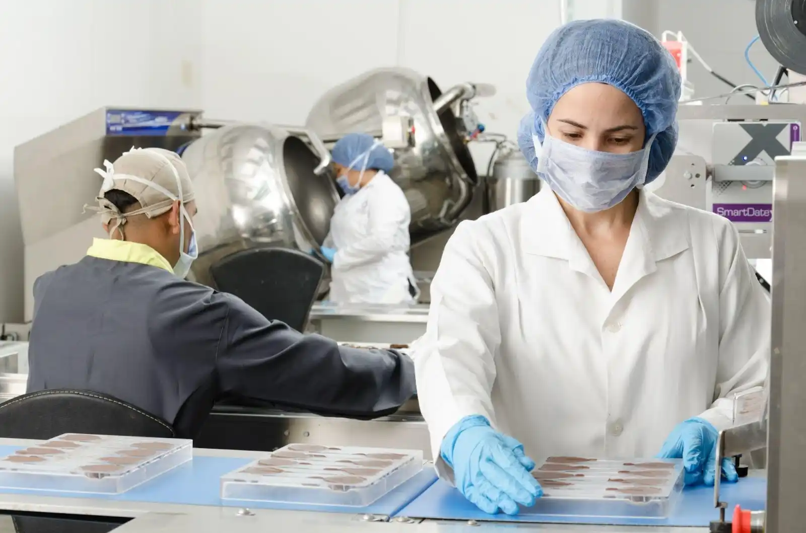 Three people in protective clothing and masks work in a food processing facility, handling trays of products near large machinery.