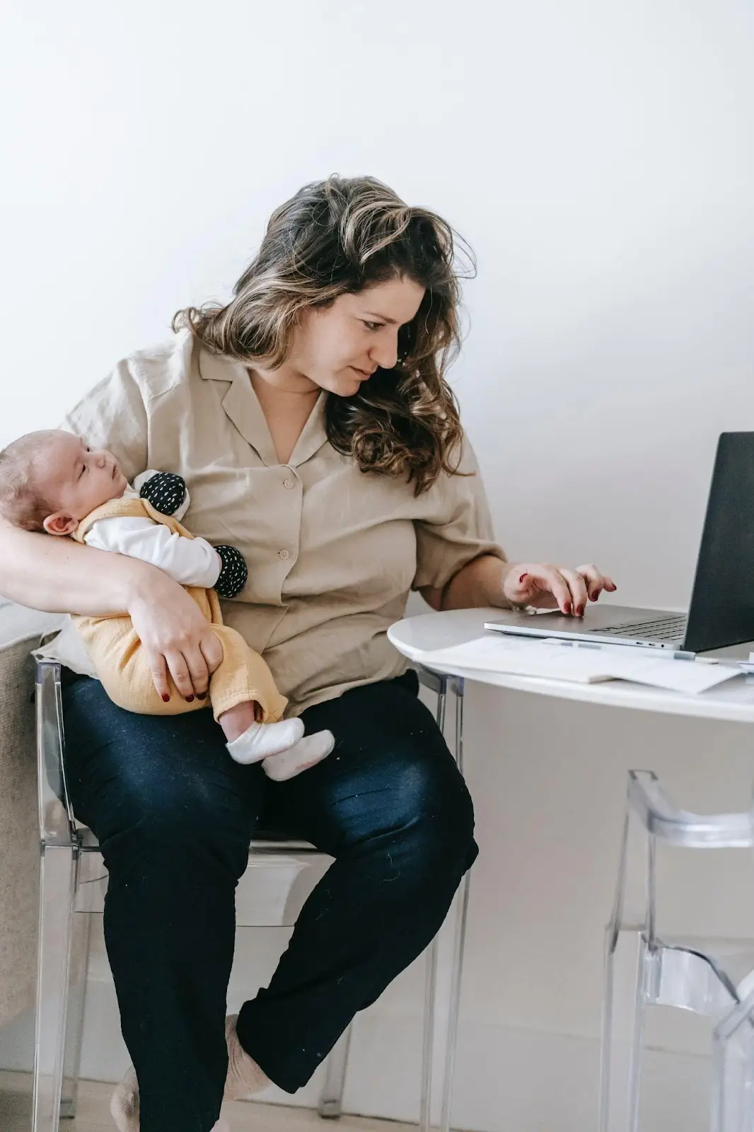 Woman balancing maternity and work, holding a baby on her lap while working on a laptop at a round table.