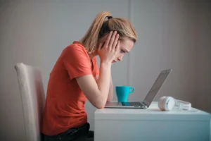 A woman in a red shirt sits at a desk, looking stressed while staring at a laptop, perhaps juggling work and maternity responsibilities. A blue mug and white headphones are on the table.