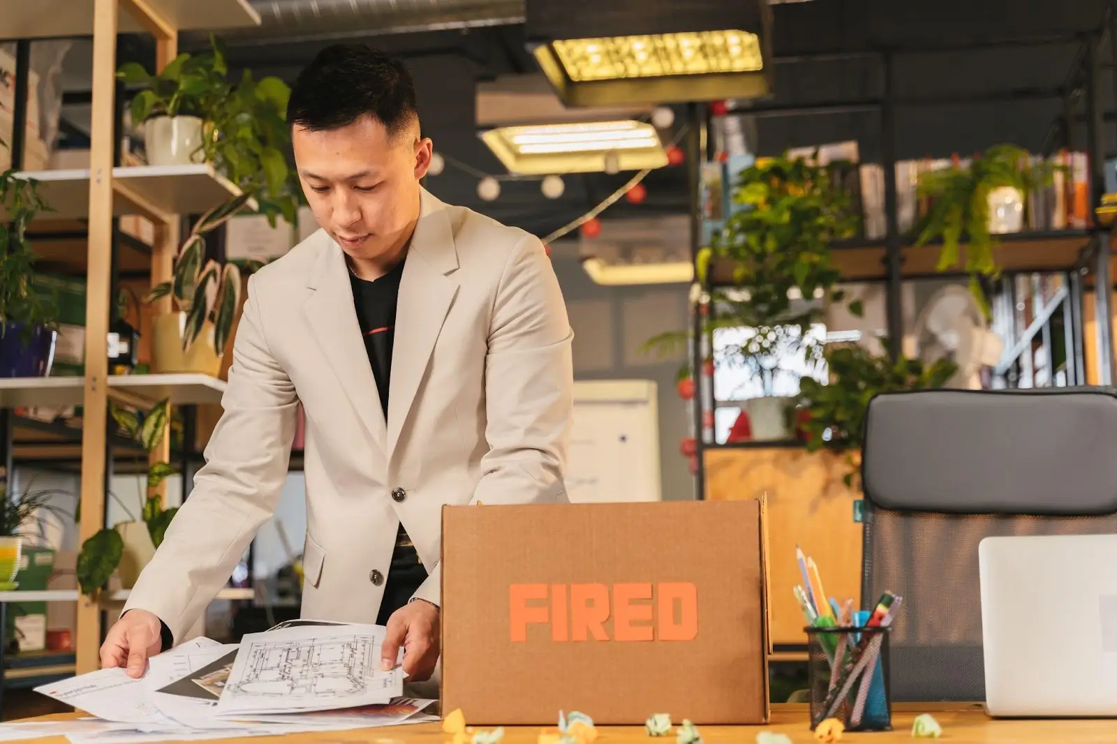 A man in a light suit gathers papers at a desk with a cardboard box labeled “FIRED” in an office filled with plants.