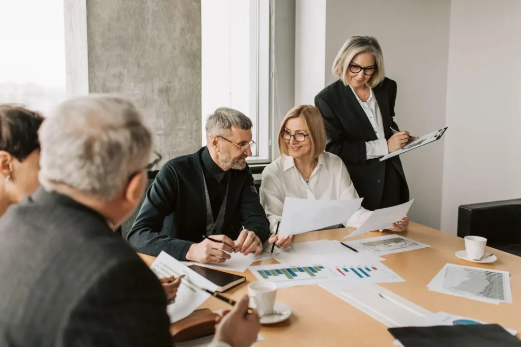Four adults in business attire sit around a conference table with documents and charts, while one person stands holding a clipboard, discussing workplace policies—including maternity considerations—in a modern office setting.