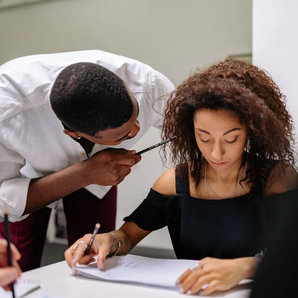 A man leans over to speak to a woman who is writing in a notebook at a desk. Both appear focused and engaged in a work or study setting.