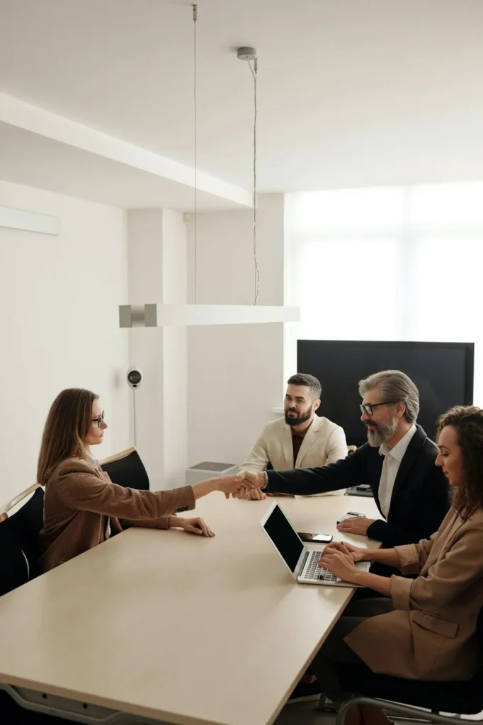 Four people in business attire sit around a conference table. One woman shakes hands with a man across the table, while two others observe, ensuring compliance with EEOC guidelines. A laptop is open on the table.