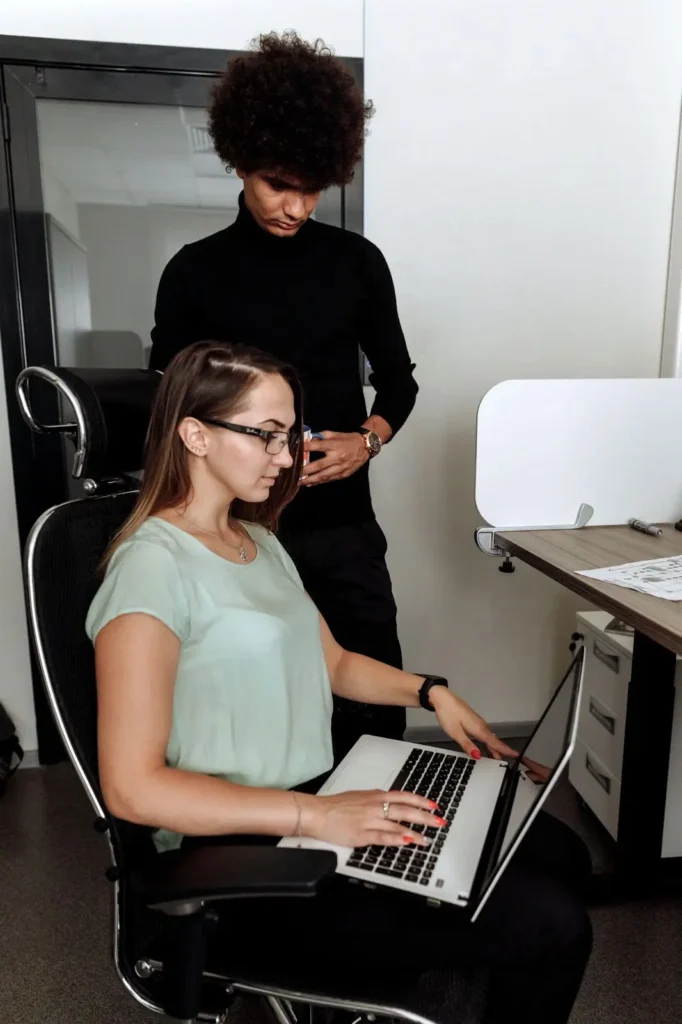 Two people in an office; one person sits working on a laptop while another stands behind, looking down at the screen.